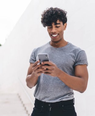 Gülümseyen, kıvırcık, gri tişörtlü genç adam beyaz duvarın önündeki merdivenlerde geziyor.