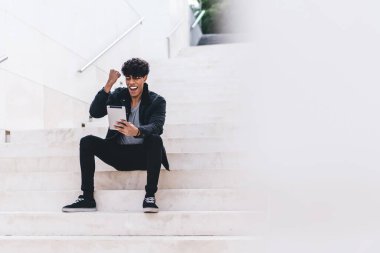 Gündüz vakti şehir merdivenlerinde otururken günlük kıyafetleriyle tablet üzerinde gezinen şık, genç, esmer, afro-amerikan bir adam.