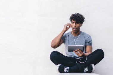 Şaşkın, genç, şaşkın, sıradan giyinen siyah erkek. Tablet ve kulaklıklarla bacak bacak bacak üstüne atarak dışarıdaki kameraya bakıyor.