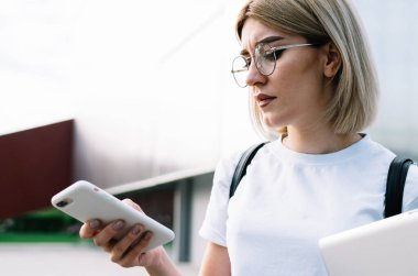 Gözlüklü, günlük giysiler içinde dikkatli bir kadın. Ofis binasının yanındaki caddede dikilirken konsantrasyonuyla akıllı telefona göz atıyor.