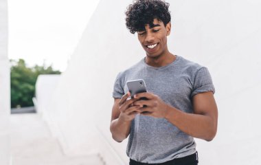 Gülümseyen, kıvırcık, gri tişörtlü genç adam beyaz duvarın önündeki merdivenlerde geziyor.