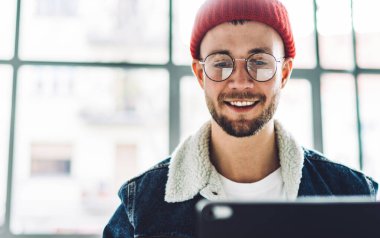 Genç, güler yüzlü, sakallı, gözlüklü, sıcak elbiseli, parlak bir odada sohbet eden, arka planda bulanık pencereli bir erkek öğrenci.