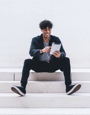 Dijital tabletli pozitif gülümseyen adam, sosyal sitede oyun oynarken elektronik kulaklıkla müzik dinlemenin keyfini çıkarıyor.