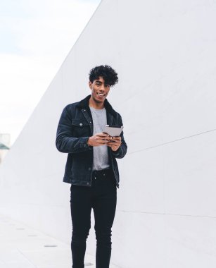 Modern stil gülümseyen pozitif esmer Afrikalı Afrikalı Amerikalı adam gün ışığında şehir caddesi tünelinde yürürken tablet üzerinde geziniyor.