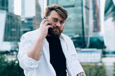 Gündelik giysiler ve yuvarlak gözlükler içinde odaklanmış bir erkek akıllı telefondan iş arkadaşıyla konuşuyor. Proje üzerinde çalışıyor. Modern şehrin sokaklarında dizüstü bilgisayarla kameraya bakıyor.