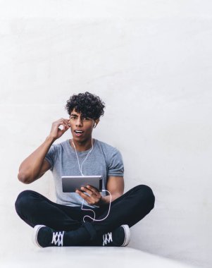 Şaşkın, genç, şaşkın, sıradan giyinen siyah erkek. Tablet ve kulaklıklarla bacak bacak bacak üstüne atarak dışarıdaki kameraya bakıyor.