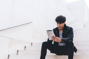 Günümüz modası geçmiş esmer, Afro-Amerikalı bir adam gündüz vakti şehir merdivenlerinde otururken tablet üzerinde geziniyor.