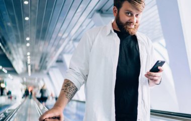 Modern cep telefonu zımbırtısıyla çevrimiçi sohbet etmek için boş zamanlarında elektrikli merdivenlerde dikilip kameraya bakan, şık giyinmiş, modern çağdan kalma hippi bir adamın yarım boy portresi.