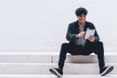 Dijital tabletli pozitif gülümseyen adam, sosyal sitede oyun oynarken elektronik kulaklıkla müzik dinlemenin keyfini çıkarıyor.