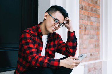 Zeki Asyalı erkek öğrenci kafaya dayanıyor ve cep telefonuyla arkadaşlarıyla mesajlaşıyor ve kapı eşiğinde kahve içerken gülümsüyor.