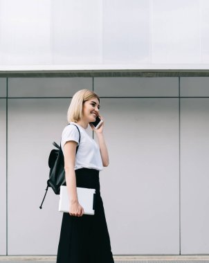 Sırtında siyah sırt çantası ve tabletle gülümseyen sarışın bir kadının sokak binasına doğru yürürken telefon etmesi.