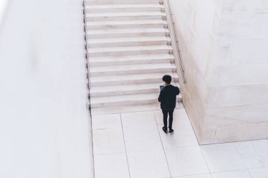 Yukarıdan siyah elbiseli bir erkeğin beyaz merdivenlerin ve duvarların yanında durduğunu ve şehirdeki tablette sörf yaptığını görüyoruz.