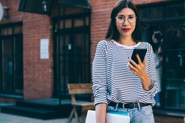 Uzak iş için dizüstü bilgisayar tutan başarılı kadın grafik tasarımcısının yarım boy portresi ve online tarama için modern hücresel cihaz ve boş zamanlarında şehir merkezinde kameraya bakmak