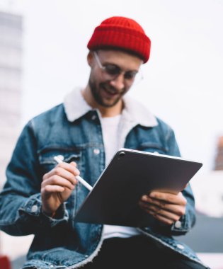 Yumuşak odaklı, güler yüzlü, gözlüklü, kırmızı şapkalı, kot ceketli, taşın üzerinde oturan ve bulanık şehir arka planında, Stylus 'la tableti tıklatan modacı genç adam.