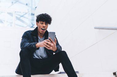 Beyaz modern binada merdivenlerde otururken kıvırcık saçlı, akıllı telefon kullanan şık giyinmiş afro-amerikalı bir adam.