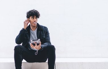 Tabletle merdivenlerde oturmuş kulaklıkla müzik dinlerken beyaz duvara bakan Afro-Amerikalı genç yakışıklı bir adam.