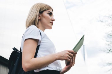 Bahçedeki binanın yanında dikilirken sırt çantalı, tablet kullanan ve dalgın dalgın bakan ciddi bir kadının yan görüntüsü.