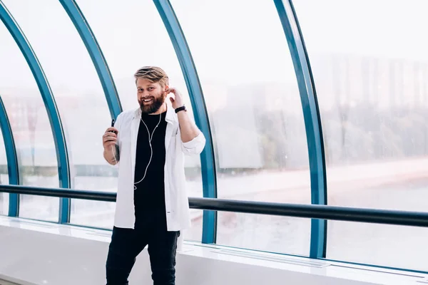 Charismatic smiling guy in white shirt with sleeves rolled up and white sneakers using mobile for listening to music adjusting earphones in pedestrian bridge