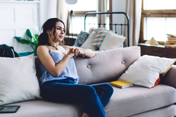 Handsome male in white T-shirt and blue jeans pointing at wristwatch and asking young sitting on couch in contemporary living room modern female to hurry up