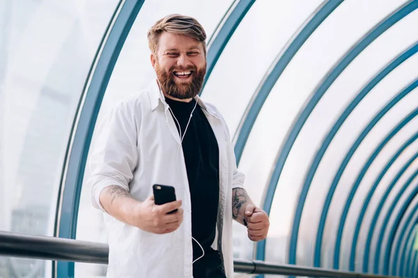 Happy laughing bearded guy in white shirt with sleeves rolled up with headphones enjoying music on smartphone in pedestrian bridge