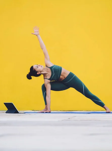 Adult slim female in tracksuit and with black hair standing on mat practicing yoga in parsvakonasana pose learning from online class while using tablet in yard on background of empty yellow wall