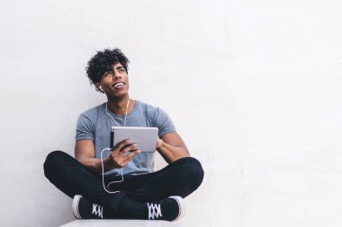 Neşeli, yakışıklı, genç, Afro-Amerikan erkek, gündelik giysiler içinde bacak bacak bacak üstüne atmış, müzik dinliyor, kulaklıklarını takıyor ve dışarı bakıyor.