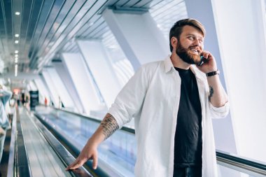 Siyah tişörtlü ve beyaz tişörtlü hoş sakallı adam cep telefonuyla konuşuyor hafif geometrik tünelde tırabzan tutuyor.