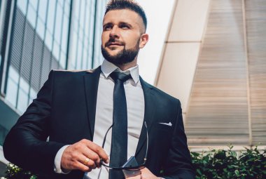 Düşük açılı, koyu renk takım elbiseli patron güneş gözlüğü ve akıllı telefon taşıyor ve şehir caddesindeki modern binaların dışındaki çalıların yanında dikilirken yüzünü çeviriyor.