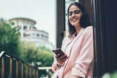 Kağıt klasörü ve günlüğü olan, cep telefonu uygulaması kullanan ve yaz günü binanın kapısında dururken gülümseyen modern başarılı bir kadından.