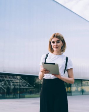 Neşeli kadın, günlük giysiler içinde, şehir caddesinde ofis binasının yanında kameraya bakarken neşe içinde tablet karıştırıyor.