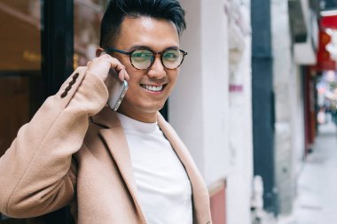 Son moda bej ceketli Asyalı erkek girişimci, ortaklarıyla cep telefonuyla konuşurken, elinde içecek ve şehirdeki sokağa bakan kameralara bakıyor.