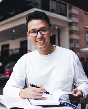 Asyalı, gözlüklü, zeki, boş not defteriyle not alan, caddedeki kafede ders çalışan ve kameraya bakan biri.