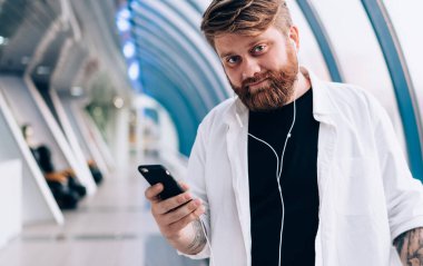 Elektronik kulaklıklı yakışıklı hippi adamın yarım boy portresi. Kapalı mekanda kameraya bakıyor ve öğrenmek için ses kitabı dinliyor. Cep telefonlu, boş zamanlarında poz veren sakallı bir adam.