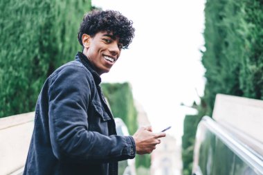 Siyah ceketli ve kot pantolonlu, yeşil ağaçlara bakan yeşil ağaçlara karşı asansörde gezen modaya uygun etnik kıvırcık erkek.