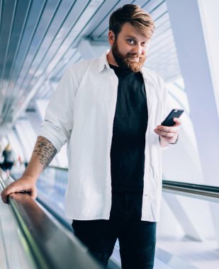 Modern cep telefonu zımbırtısıyla çevrimiçi sohbet etmek için boş zamanlarında elektrikli merdivenlerde dikilip kameraya bakan, şık giyinmiş, modern çağdan kalma hippi bir adamın yarım boy portresi.