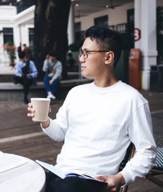 Asyalı, düşünceli, gözlüklü genç bir erkek öğrenci elinde kahve ve kitapla şehir kafesinde oturuyor ve gözlerini kaçırıyor.