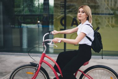 Kendine güvenen, gündelik kıyafetli, siyah sırt çantalı, bisiklet süren ve kameraya bakan modern sarışın kadının yan görüntüsü.