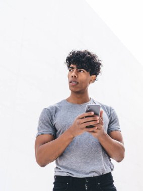 Düşünceli Afro-Amerikalı genç bir erkekten cep telefonuyla sörf yapan ve açık havada bulanık beyaz arka plana bakan.