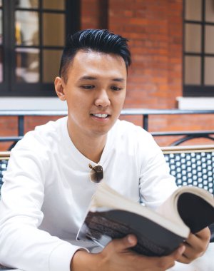 Beyaz tişörtlü yakışıklı Asyalı bir erkek kahve fincanı ile masada oturuyor ve kafede vakit geçirirken yeni bir kitap çeviriyor.