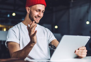 Kırmızı bereli genç adam oturmuş tabletle sohbet ediyor. Geniş endüstri ofisindeki masada çalışırken video görüşmesi yapıyor.