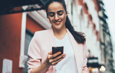 Gözlüklü, siyah saçlı, gözlüklü, şık elbiseli, beyaz perdeye konsantre olan ve cadde boyunca bulanık cephelere karşı yürürken akıllı telefonla etkileşim kuran bir kadın.