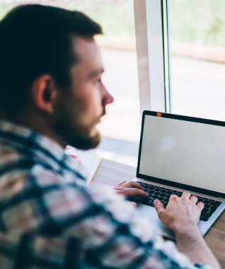 Yoğunlaşmış, koyu renk saçlı, günlük gömlekli, kahve dükkanındaki masada oturan dizüstü bilgisayarla yeni projenin planını yazan adam.