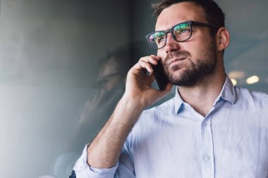 Mavi gömlekli ve mavi pantolonlu ciddi sorunlu bir erkek camdan kapının karşısındaki ofiste cep telefonundan arama yapıyor.
