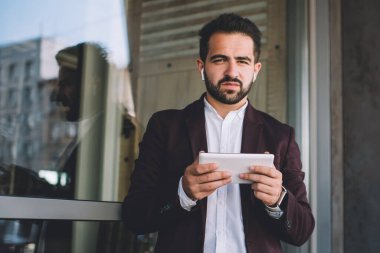 Takım elbiseli asık suratlı etnik adam modern binanın penceresinin yanındaki tableti incelerken kameraya bakıyor ve müzik dinliyor.