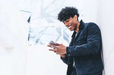 Beyaz modern binada dikilirken, kıvırcık saçlı, akıllı telefonuyla mesajlaşan şık giyinmiş neşeli, Afro-Amerikan bir adam.