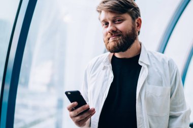 Yakışıklı hipster blog yazarının yarım boy portresi, günlük beyaz gömlekli cep telefonu cihazını elinde tutuyor ve boş zamanlarında kameraya bakıyor, teknoloji ve iletişim kavramı.