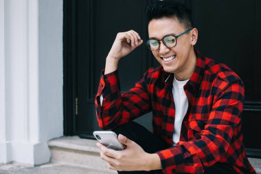 Neşeli genç Asyalı erkek öğrenci, günlük kıyafetler içinde cep telefonu kullanıyor ve bina kapısında otururken eğleniyor.