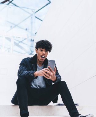 Beyaz modern binada merdivenlerde otururken kıvırcık saçlı, akıllı telefon kullanan şık giyinmiş afro-amerikalı bir adam.