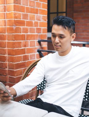 Sokak kahvecisindeki masada oturup dinlenirken telefonu kullanan, gündelik kıyafetlerle tatmin olmuş genç bir erkek.