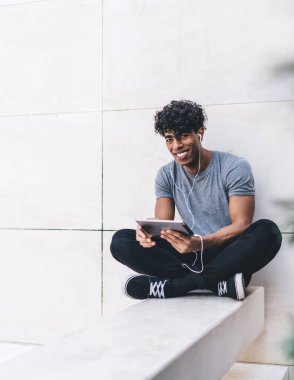 Neşeli, mutlu, yakışıklı, gündelik giyinmiş siyah erkek. Tablet ve kulaklıklarla bacak bacak bacak üstüne atmış ve dışarıdaki kameraya bakıyor.